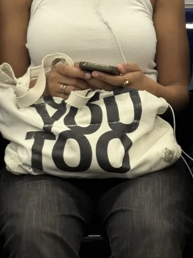 A person with a tote bag that reads "You too", looking at their phone