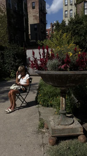 A woman sitting on a folding chair in a small garden beside a stone planter
