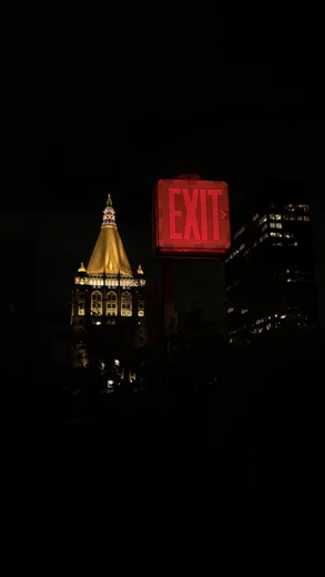 A dark night skyline with a lit spire and a red exit sign