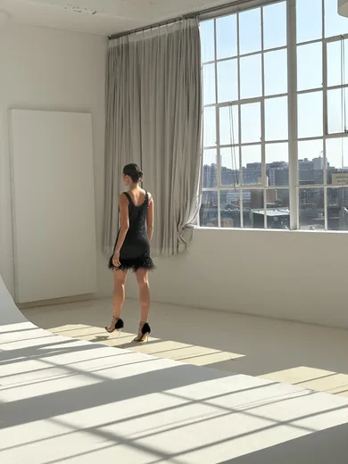 A woman in a black dress walking through a sunlit white room with large windows